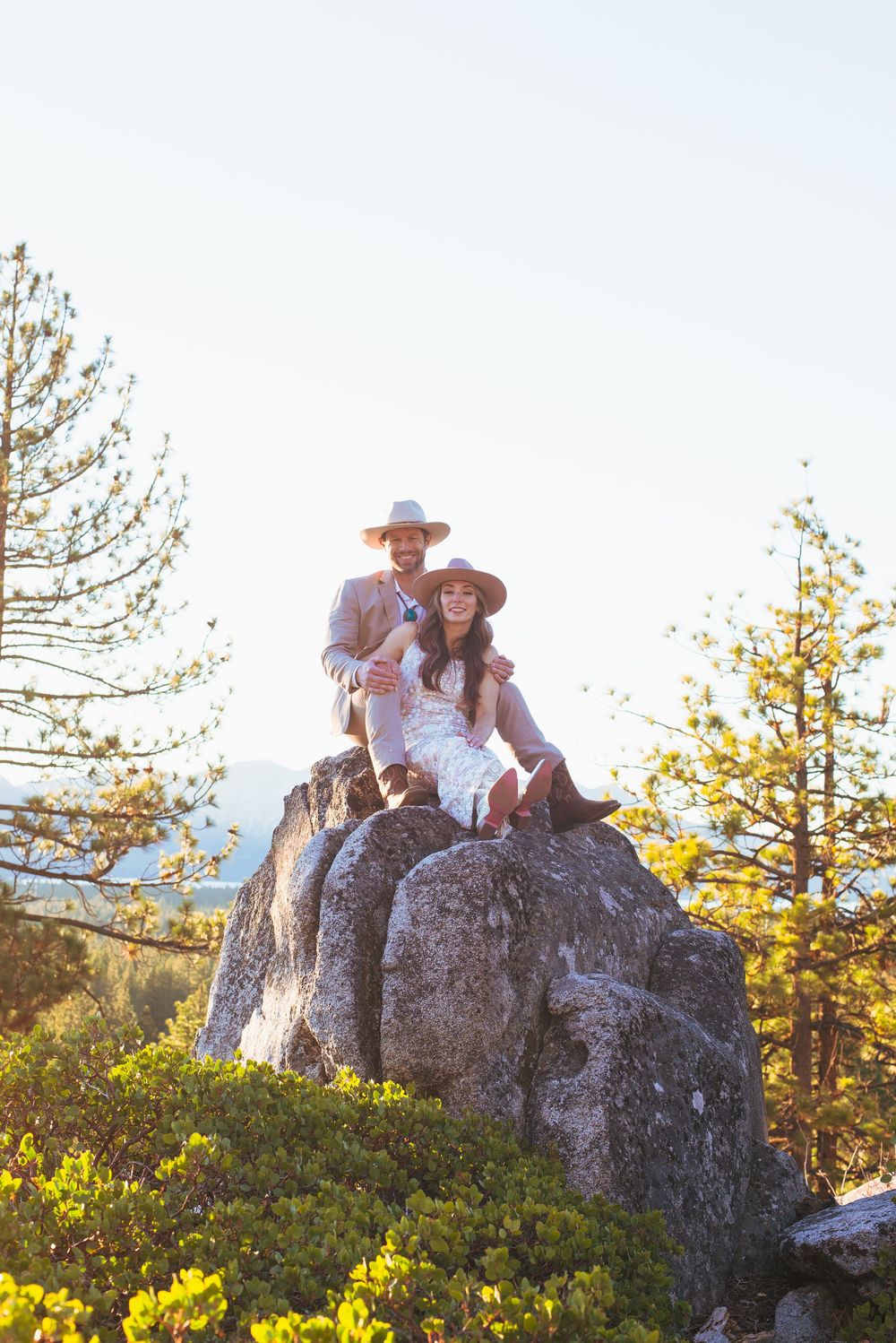 Bride and groom in cowboy hats sitting together on large boulder surrounded by pines.