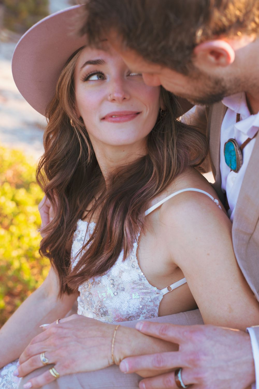 Close-up of smiling bride in pink hat being embraced by groom at outdoor wedding.