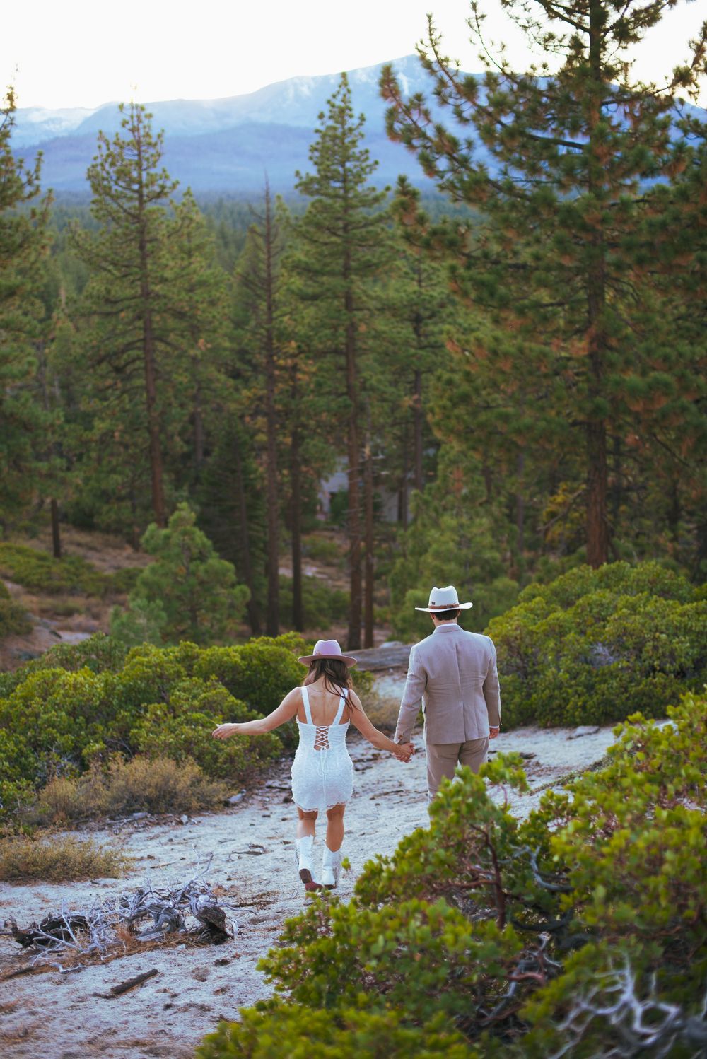 Couple holding hands walking through pine forest trail in wedding attire with cowboy hats and mountain views.