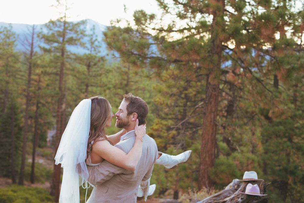 Groom lifting bride in wedding veil among pine trees with mountain backdrop at sunset.