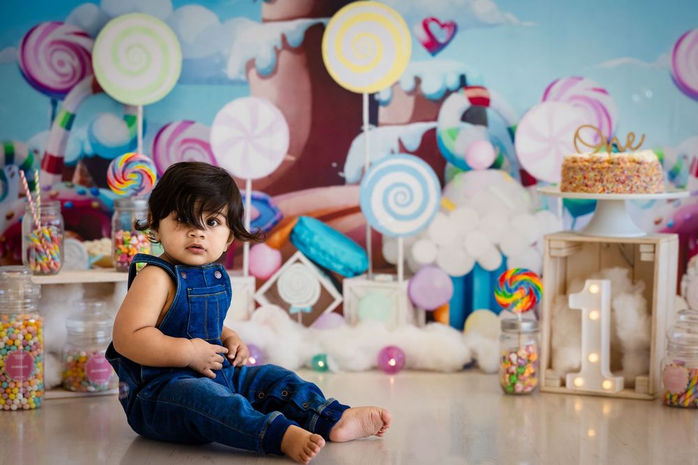 Baby in denim overalls sits on floor during colorful candy-themed first birthday cake smash photo session.