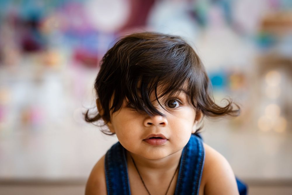 Adorable toddler with dark hair and big brown eyes wearing a blue top, looking curiously at the camera.