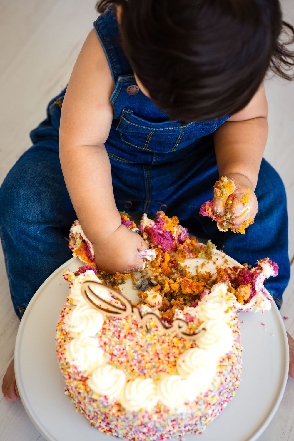 Baby in denim overalls smashing a colorful sprinkle birthday cake, grabbing handfuls of frosting and cake.
