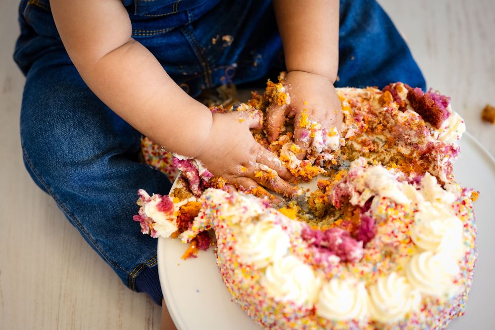 Baby in denim overalls smashing a colorful birthday cake with both hands during a cake smash photoshoot.
