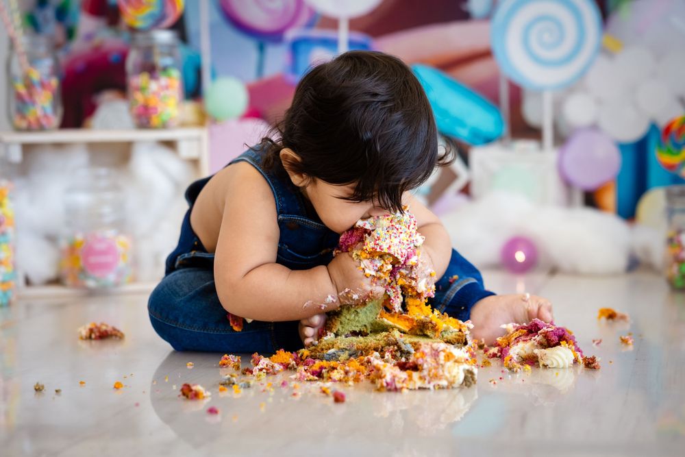 Toddler smashing colorful birthday cake on floor during cake smash photoshoot with candy backdrop.