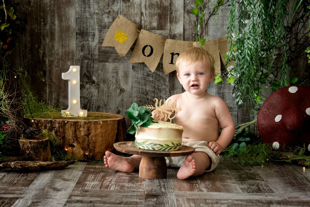 Baby's first birthday cake smash photo with woodland theme decor, number one prop, and burlap banner on rustic wood backdrop.