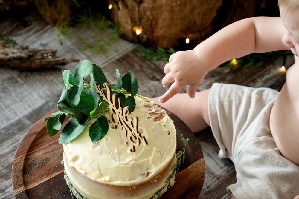 Baby reaching into a rustic first birthday smash cake decorated with greenery on a wooden board.