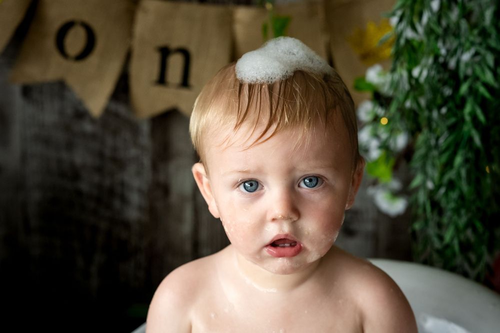 Cute baby with blue eyes and soap suds on head sitting in bath tub celebrating first birthday.