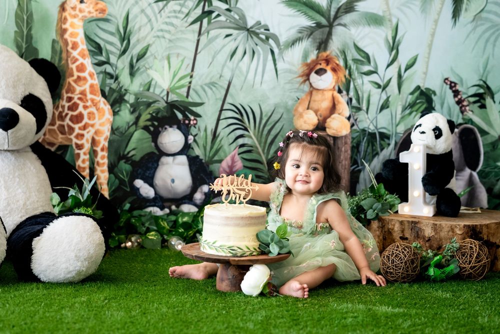 Toddler girl in green dress sits with birthday cake surrounded by stuffed jungle animals on artificial grass.