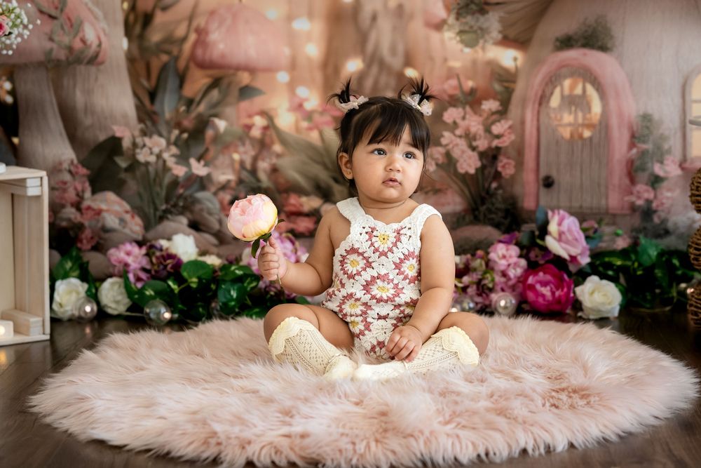 Baby girl in floral romper sits on pink fluffy rug surrounded by flowers and fairy tale decorations.