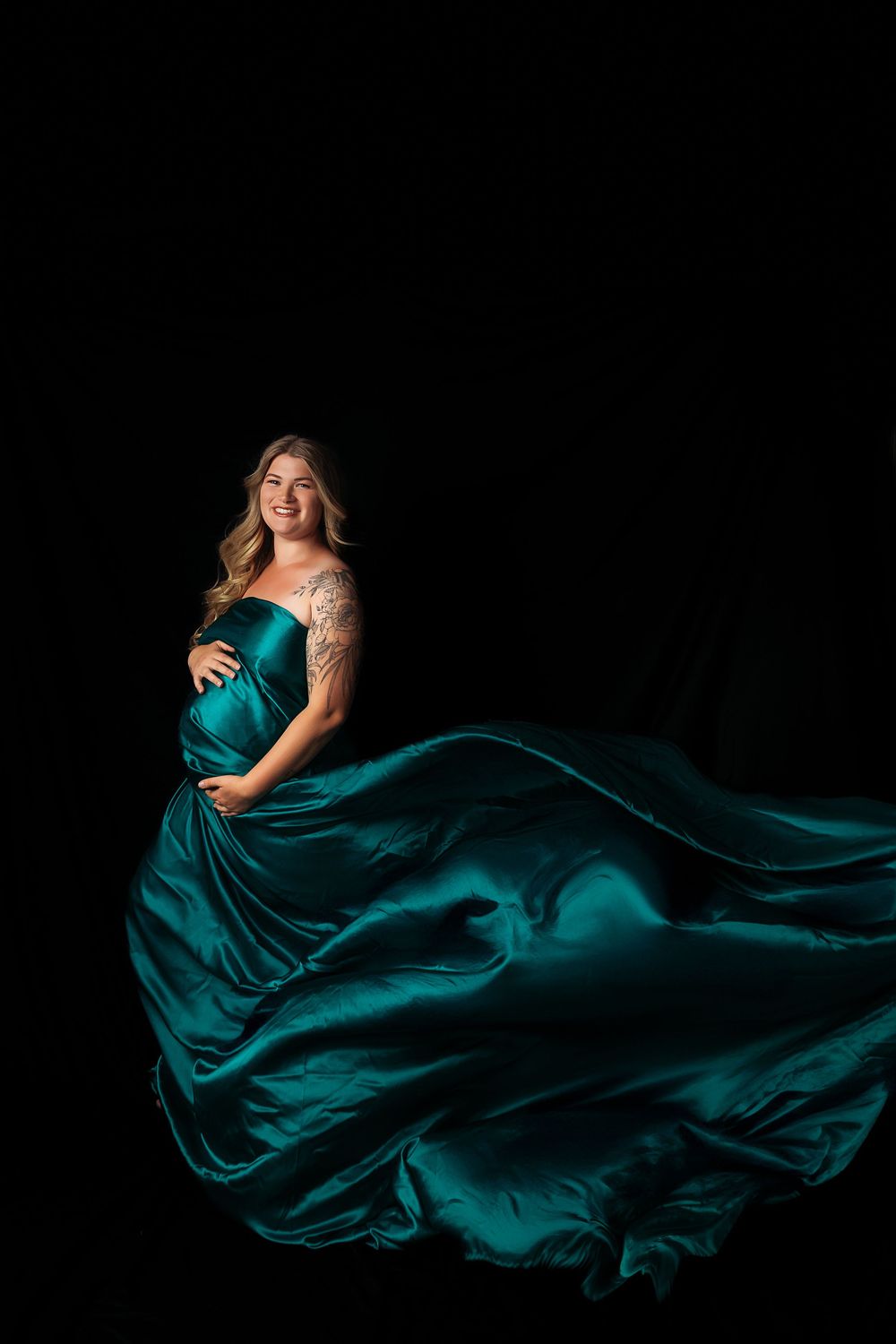 Dramatic maternity photos in flowing teal silk dress against black background with fabric motion.