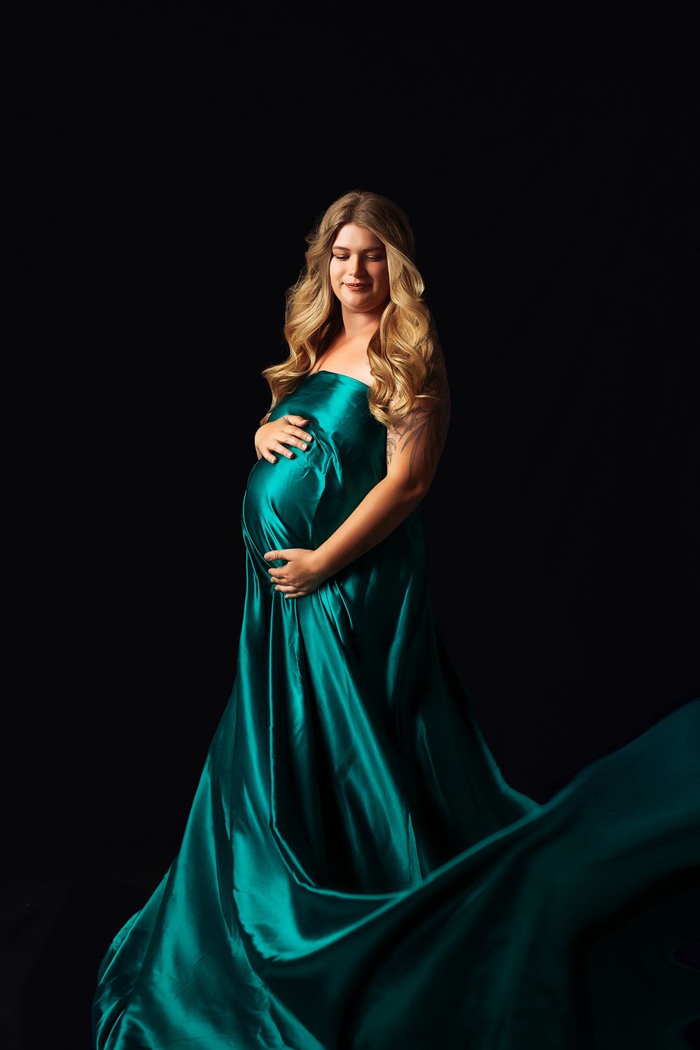 Elegant maternity portraits against black background with flowing emerald green dress creating dramatic movement.