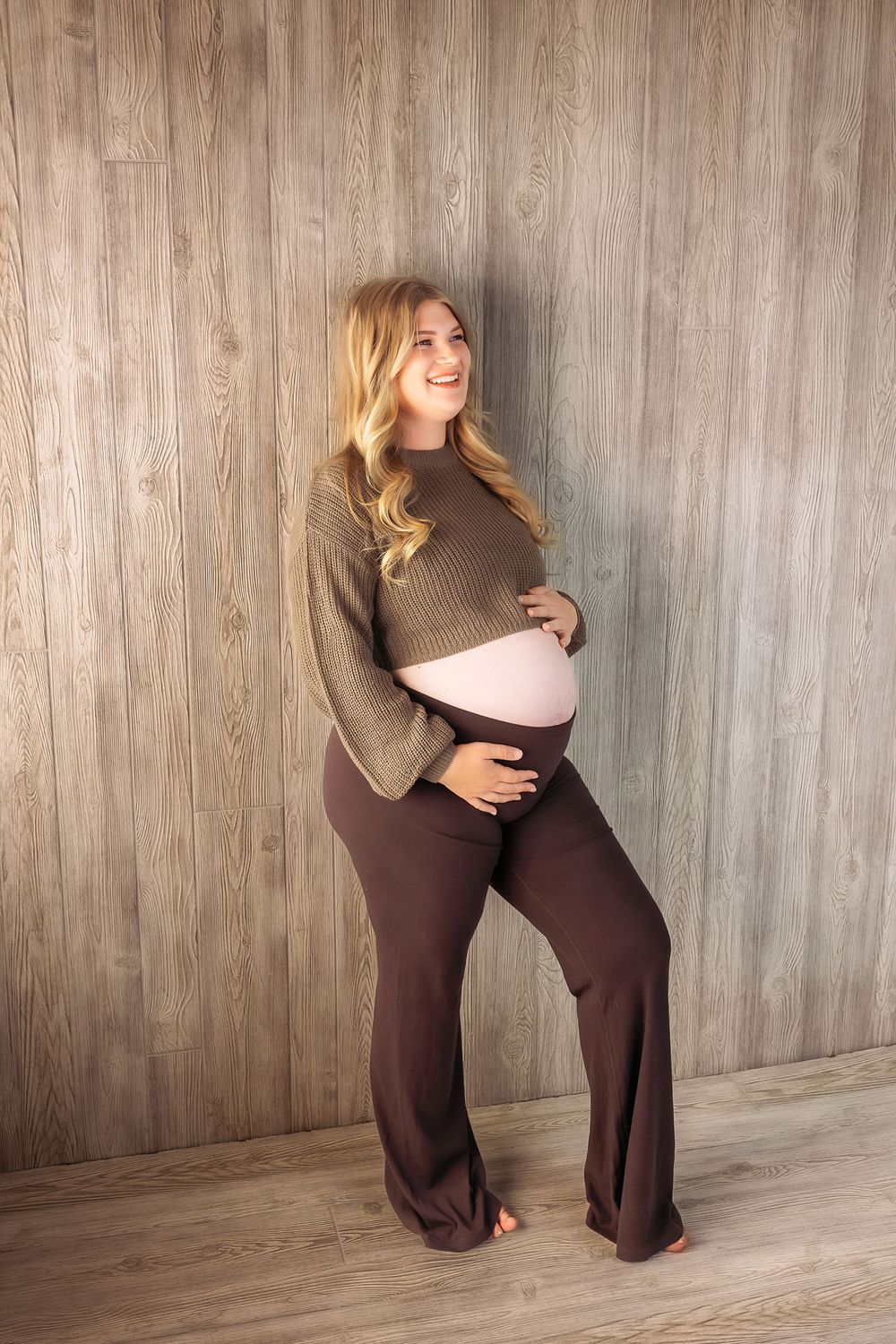 A maternity photoshoot sequence showing poses in brown pants and sweater against a wooden wall.
