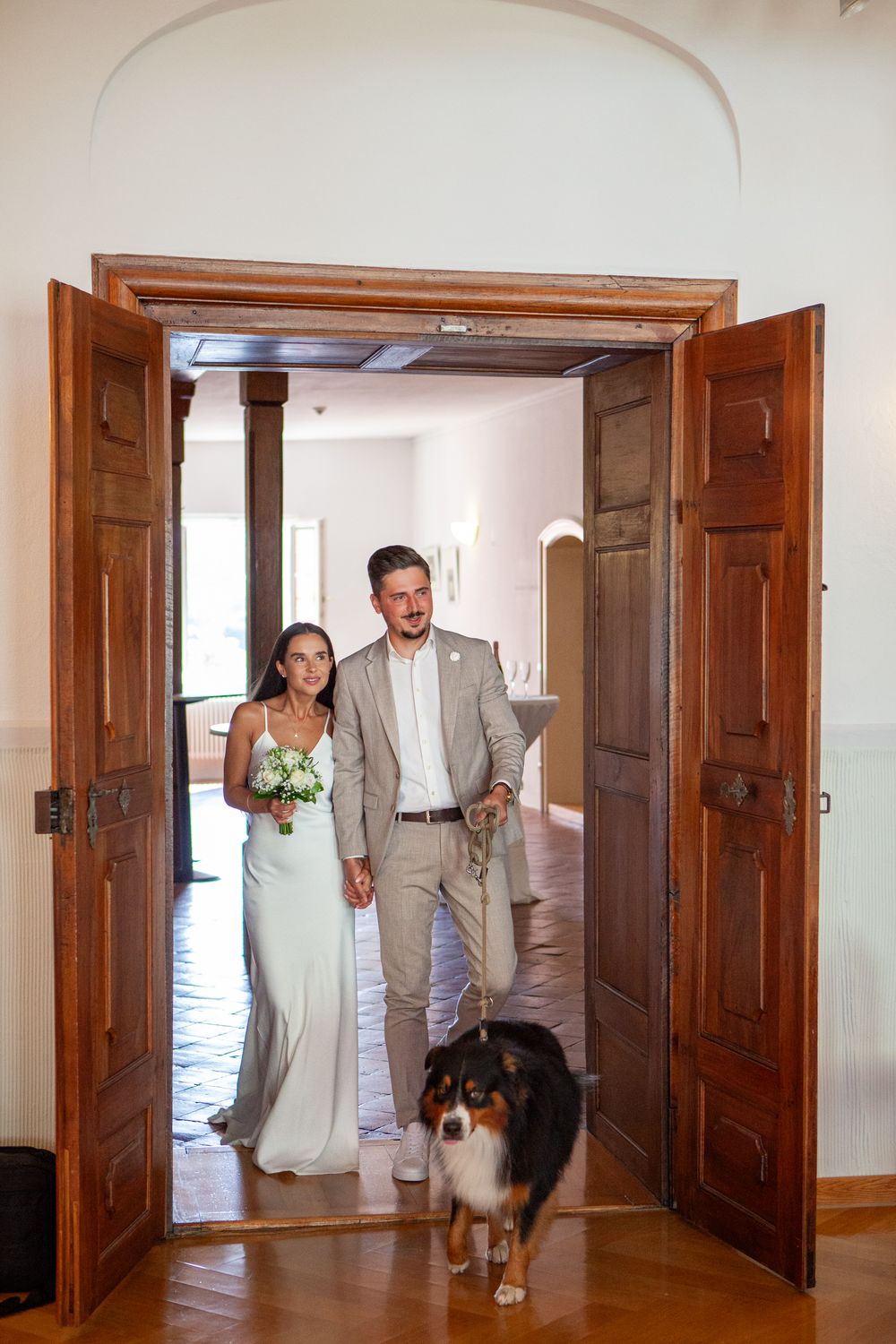 A dog follows people through a hallway with wooden doors and white walls during a wedding event.