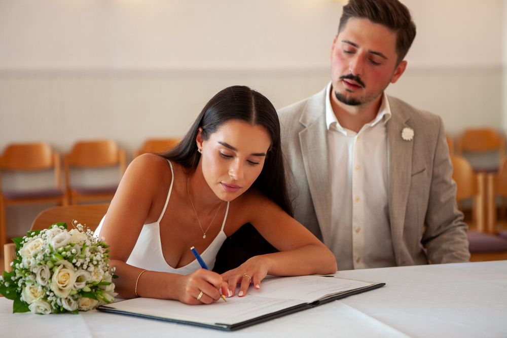 A couple signs marriage documents at a table while others observe the ceremony.