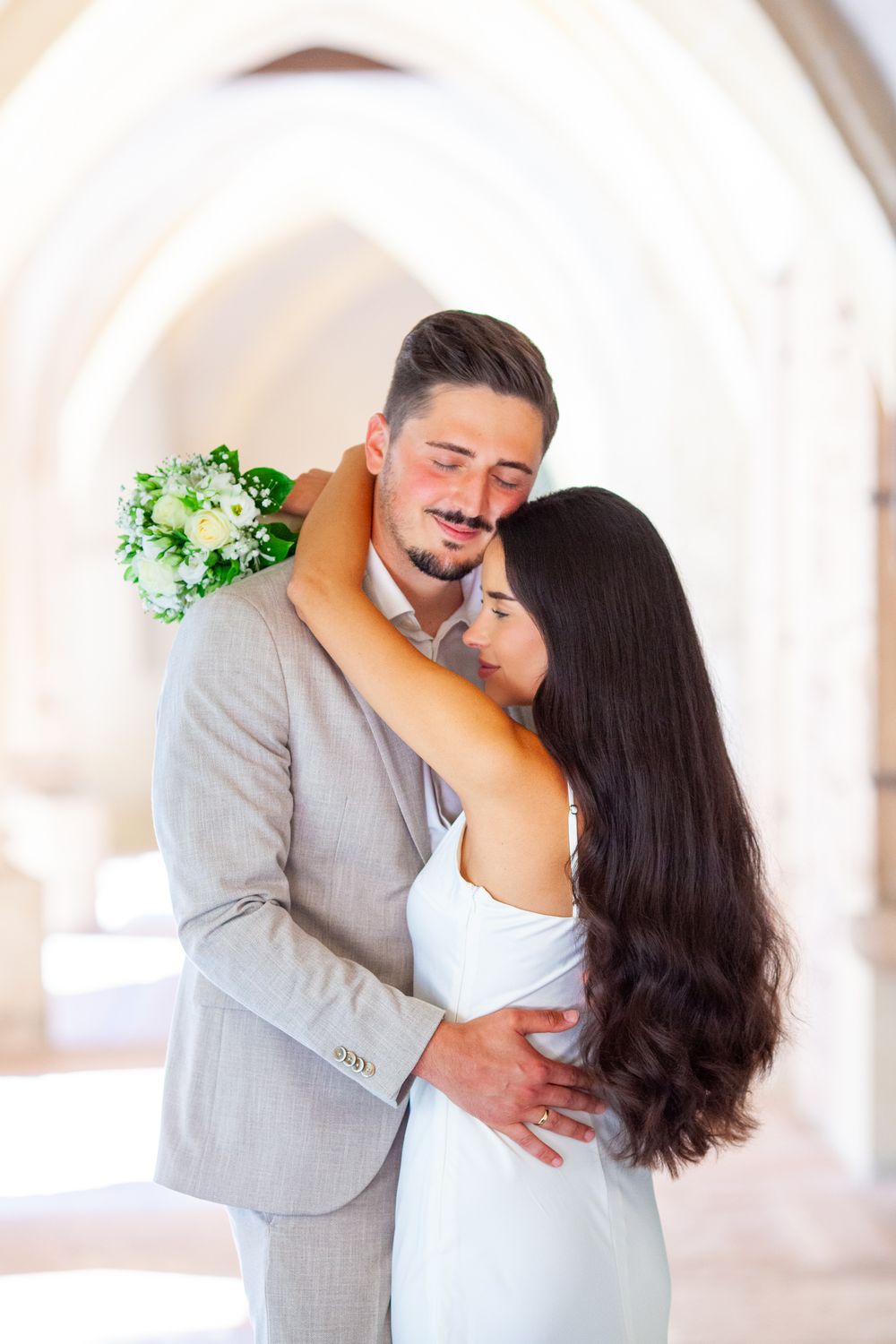 A romantic couple in light colored attire share an intimate dance pose in a bright white room with greenery accents.
