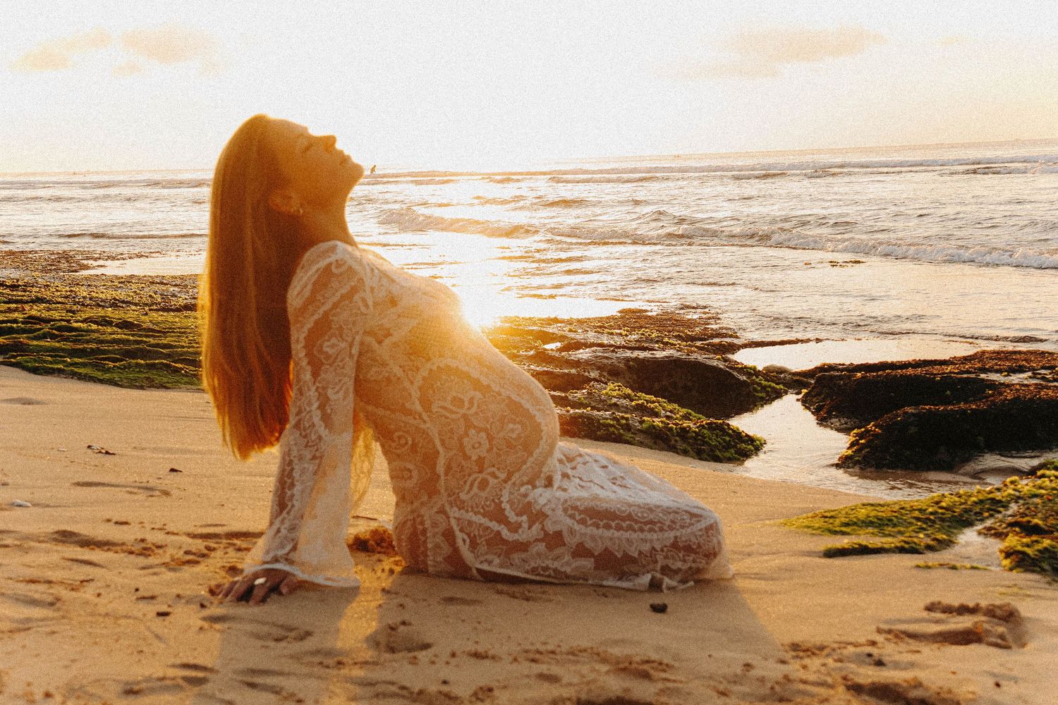 Pregnant woman in lace dress sitting on beach at glowing golden sunset.