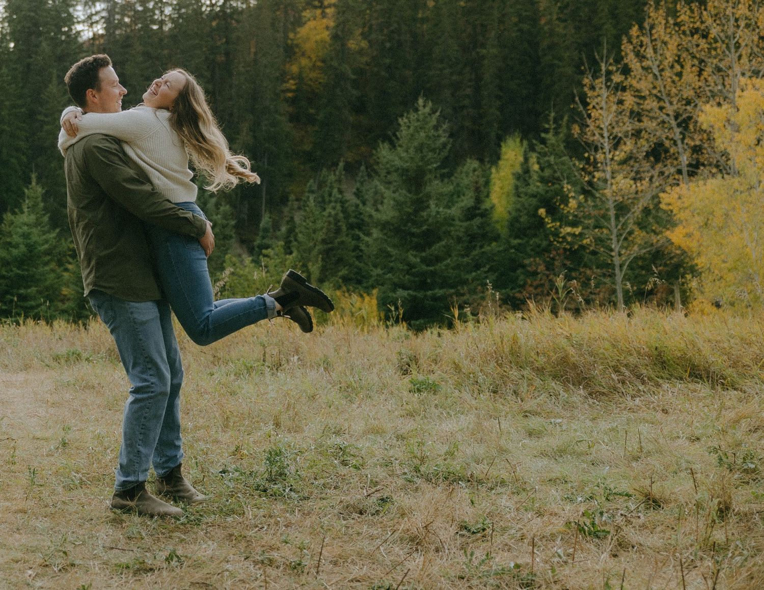 Couple in casual attire embraces while standing in a grassy meadow with autumn trees in the background.