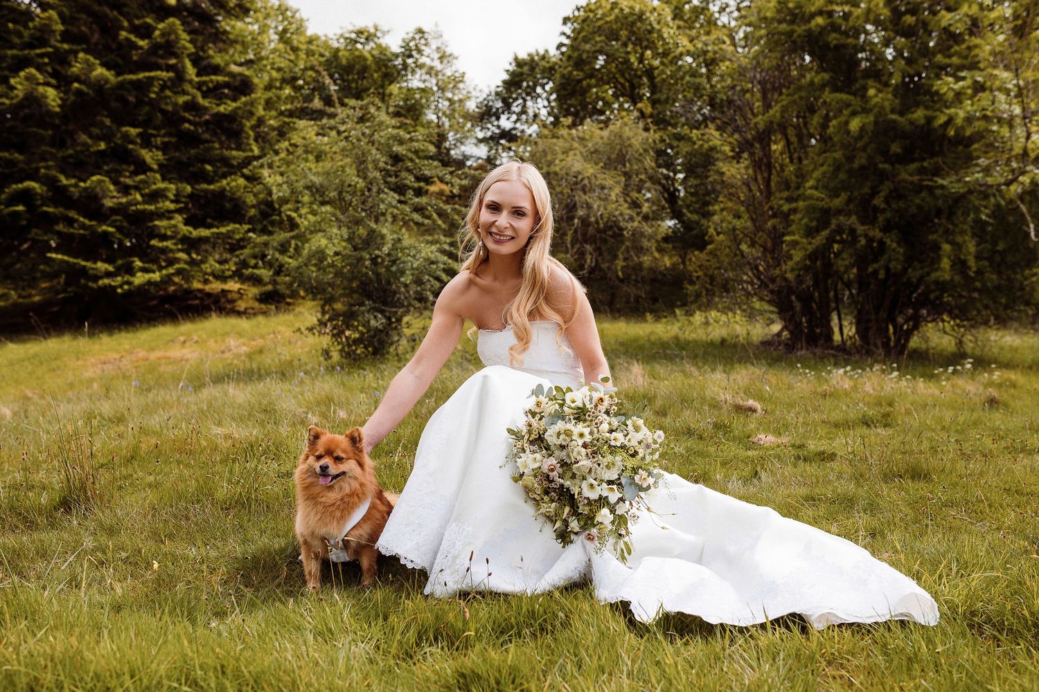 A bride in a white wedding dress sits gracefully in a sunlit meadow surrounded by lush green grass and trees.