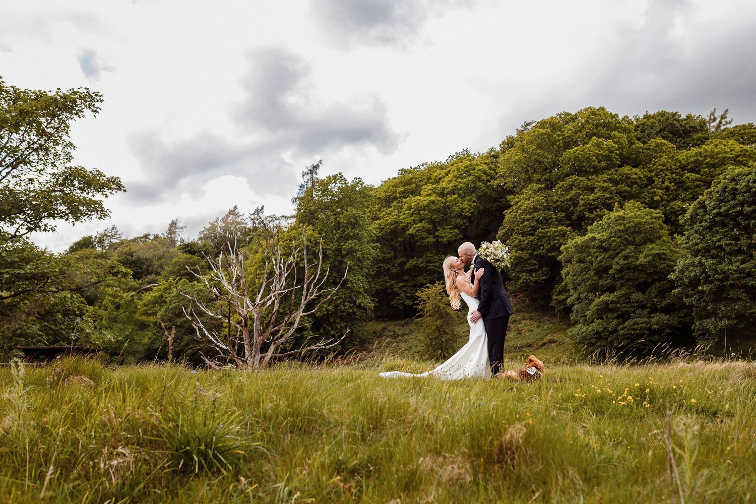 A couple shares a romantic moment in a scenic meadow surrounded by lush green trees and overcast skies.
