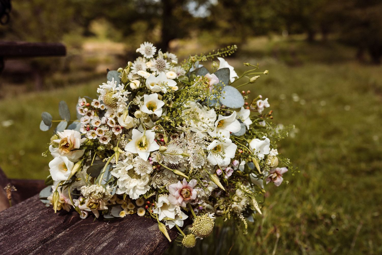 A rustic wedding bouquet with white flowers and greenery rests on a wooden surface in an outdoor setting.