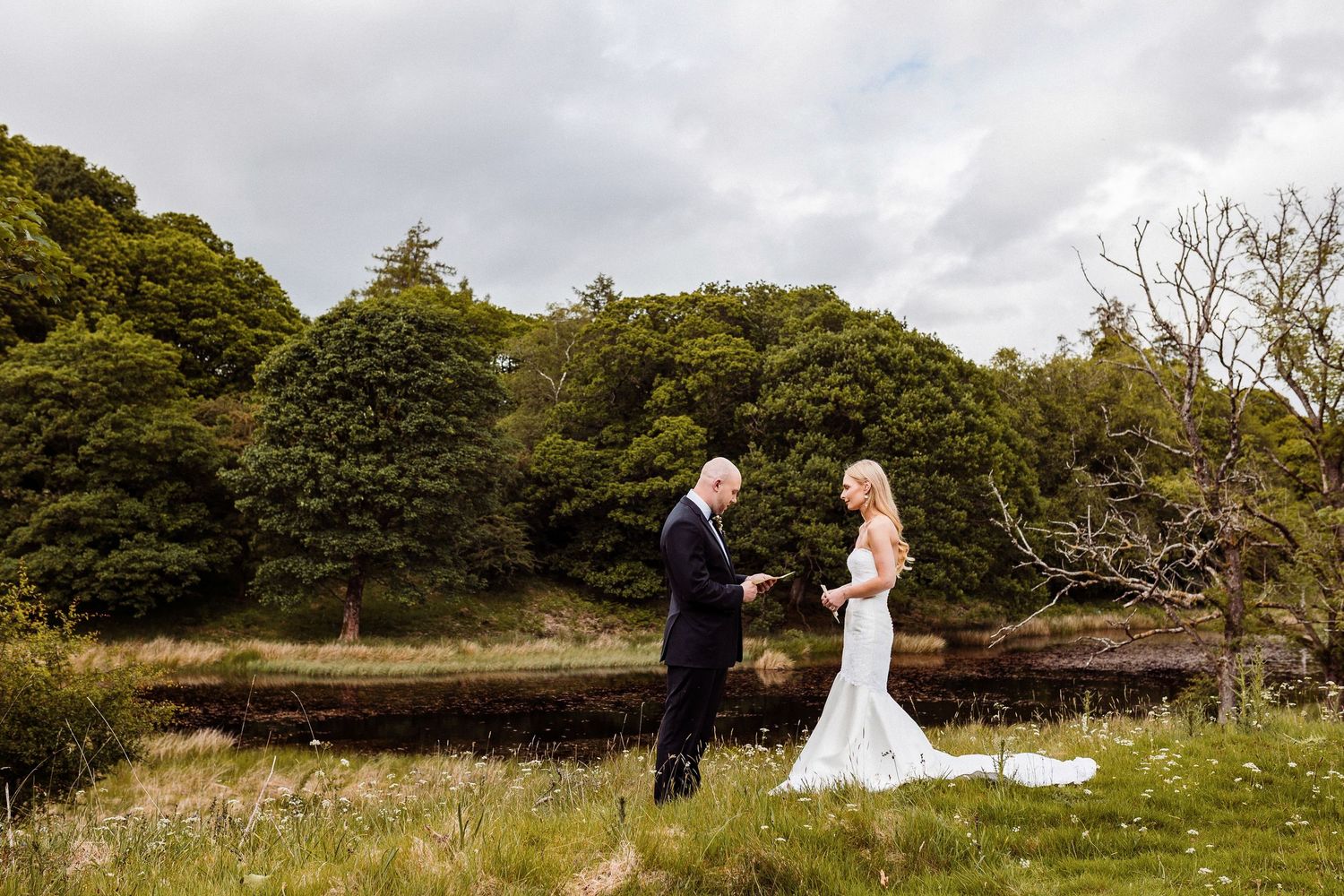 A couple shares a romantic moment in their wedding attire against a backdrop of lush green trees and cloudy skies.