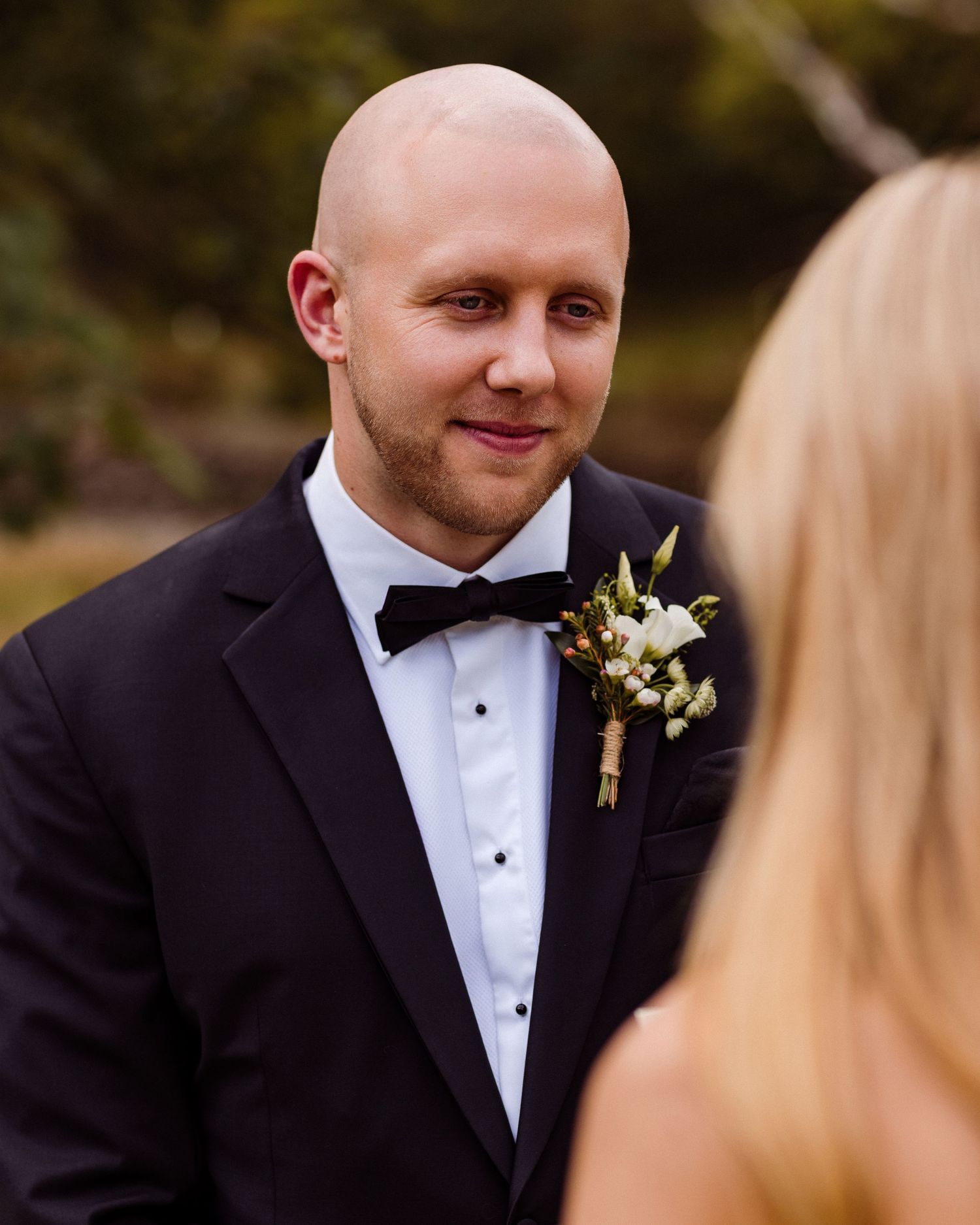 A groom in a black tuxedo and bowtie smiles during an outdoor wedding ceremony.