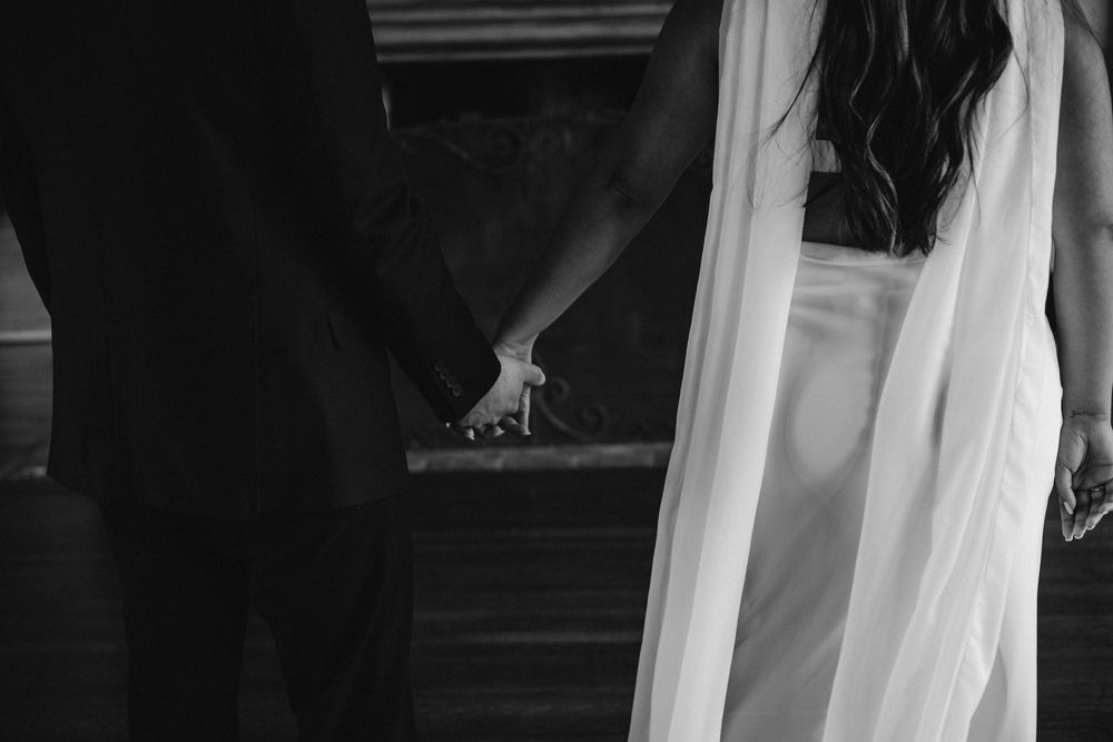 Black and white close-up of newlyweds holding hands during wedding ceremony with flowing white veil visible.