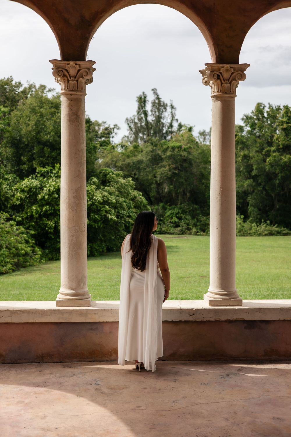 A person in a flowing white dress stands between ornate stone columns overlooking a lush green landscape.