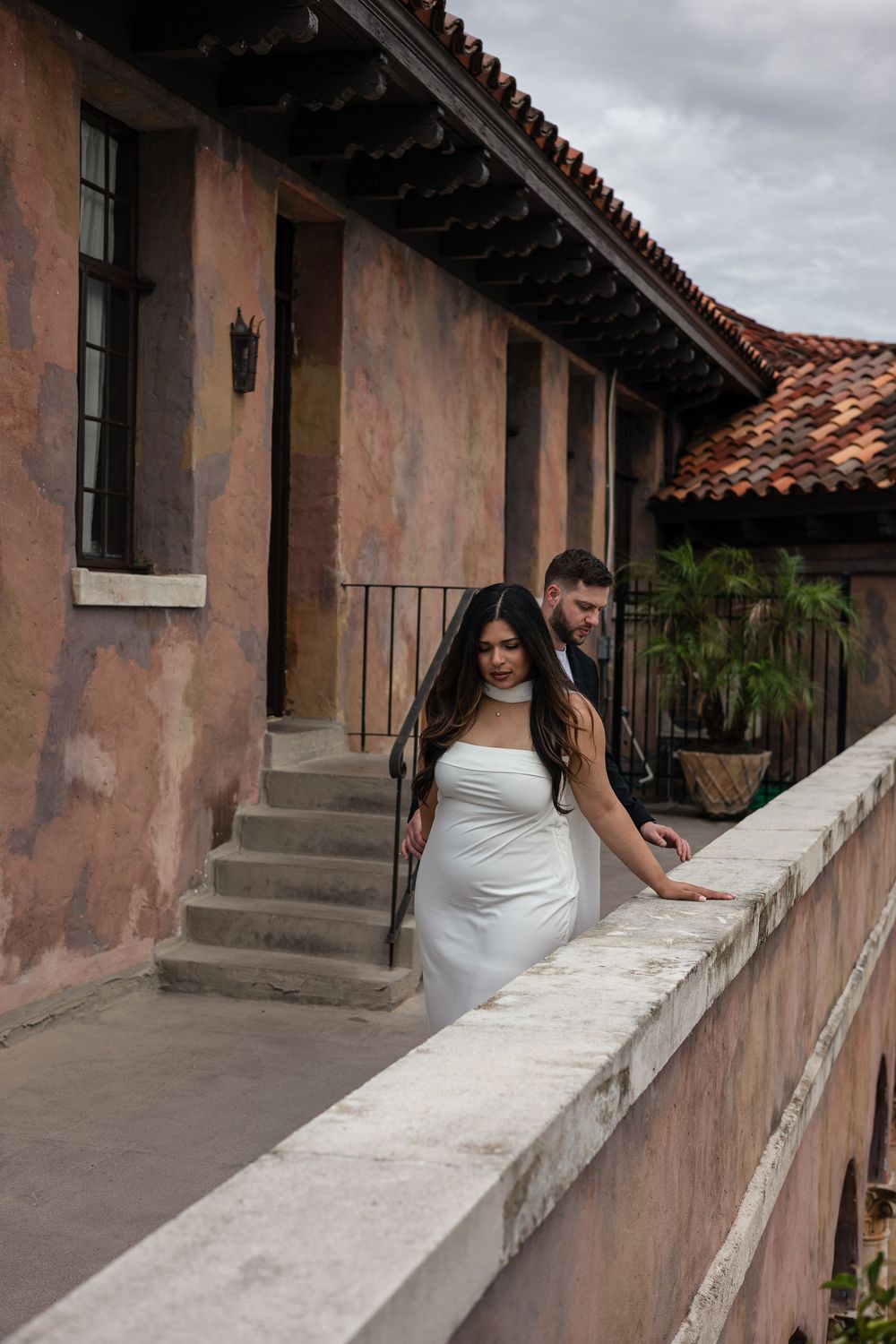 People stand along a covered walkway with terracotta roof and pink stucco walls at a historic Spanish colonial building.