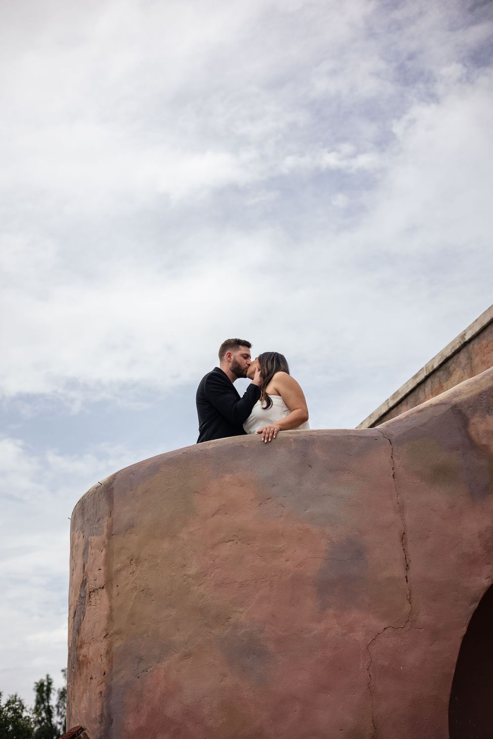 A couple sits intimately atop a large red rock formation against a cloudy sky.