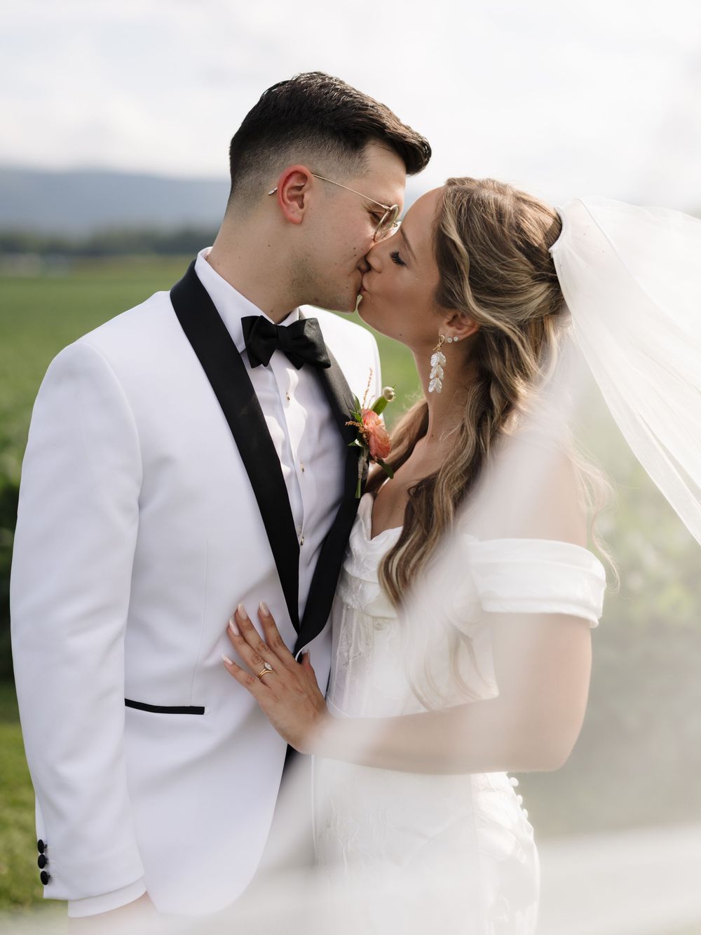 Romantic wedding couple sharing intimate kisses in white formal attire with flowing veil against scenic outdoor backdrop.