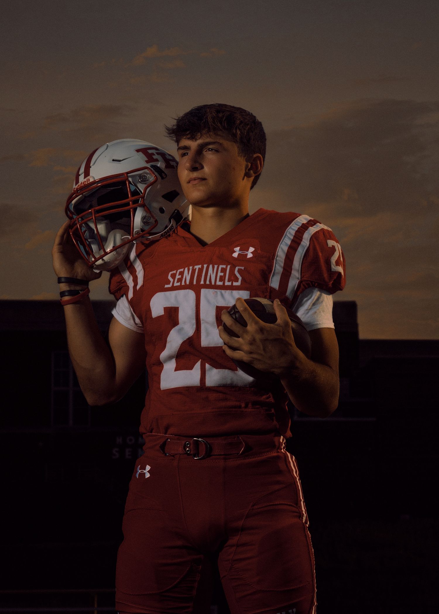 Football player in red Sentinels uniform #25 poses with helmet against sunset sky.