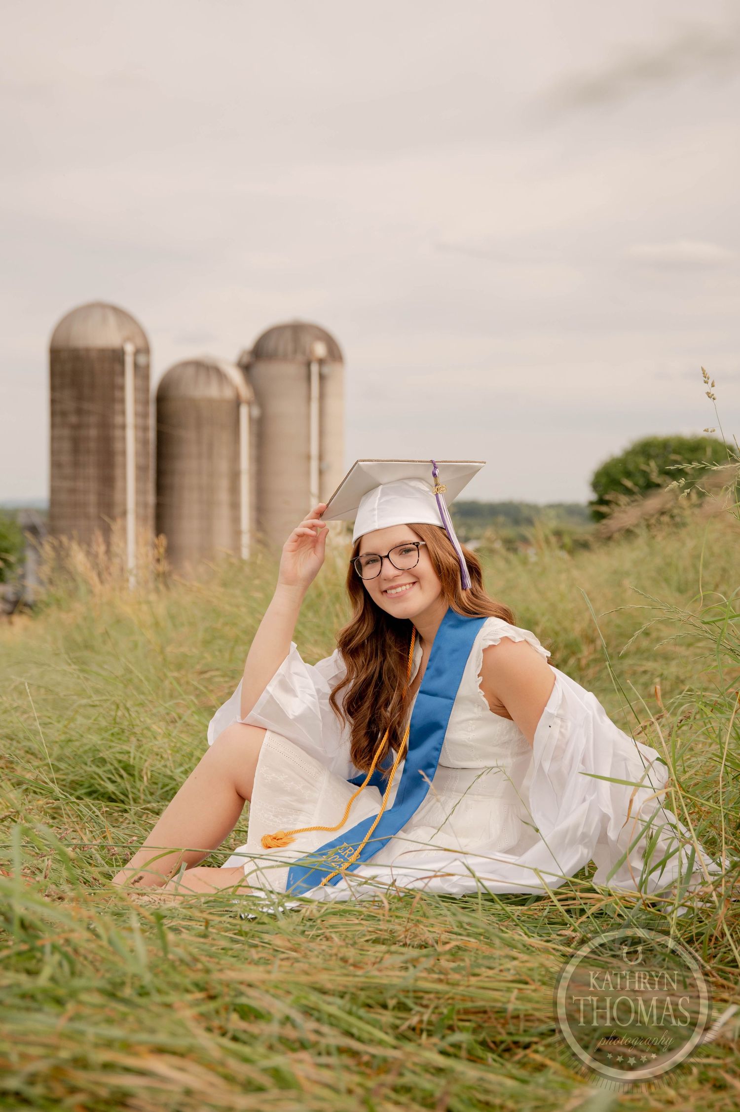 A person in a white dress and blue sash reclines in a grassy field with silos visible in the background.