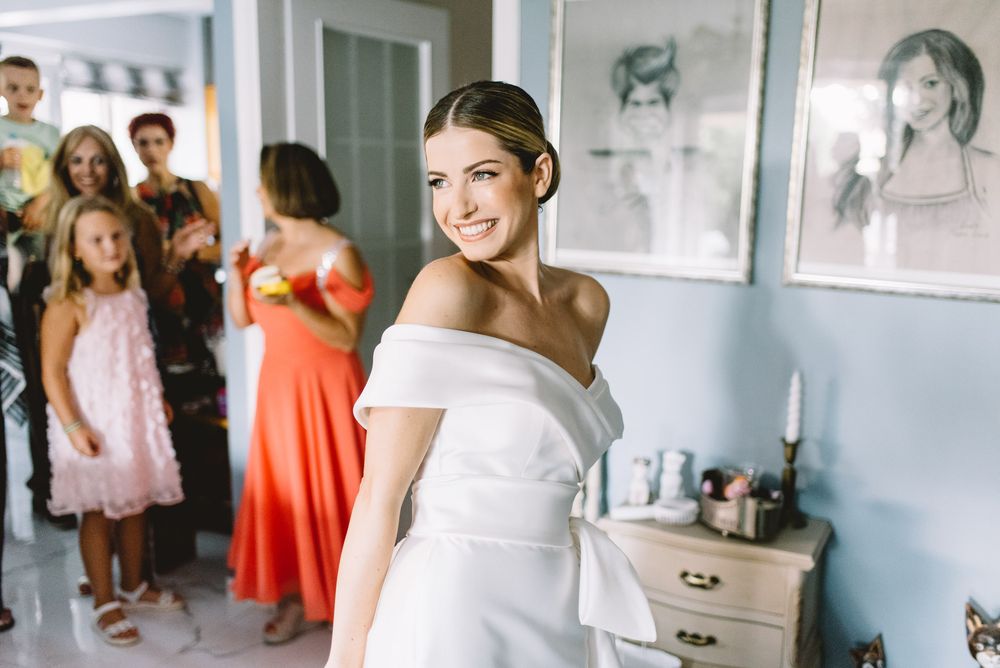 A radiant bride wearing an elegant off-shoulder white gown stands in a bright room while guests gather in the background.
