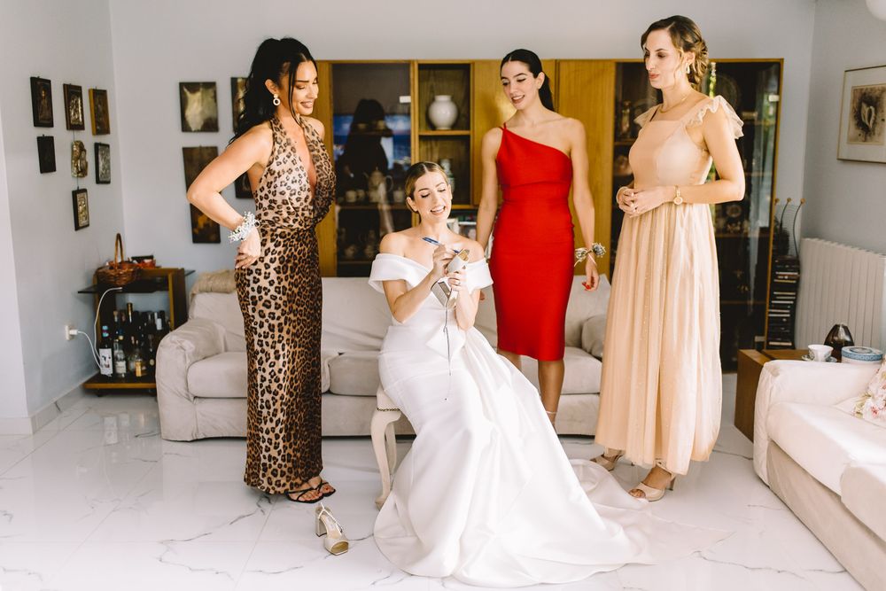 Four women share a joyful candid moment in formal dresses including leopard print, red, and white gowns in a living room.
