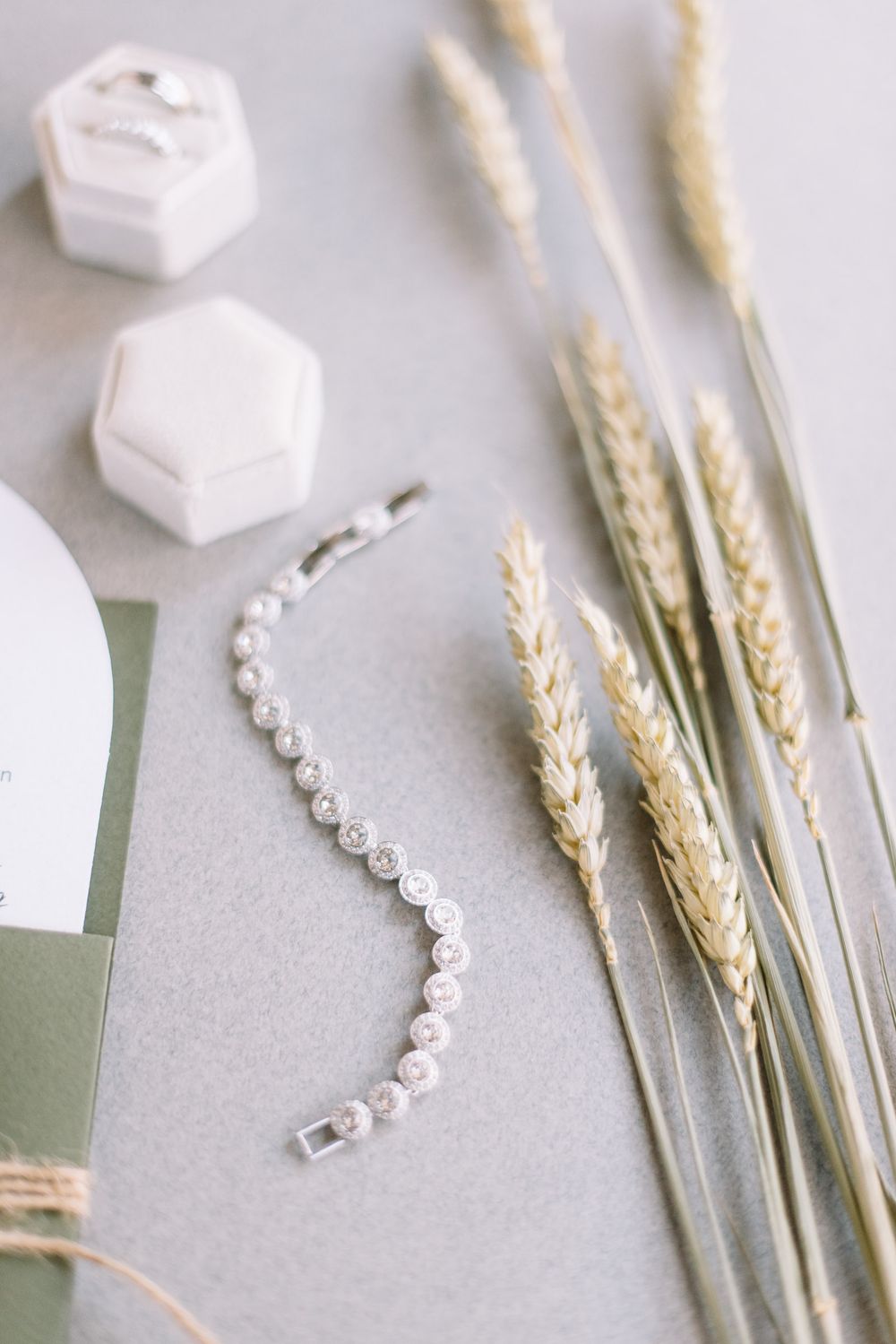 Wedding rings and delicate dried wheat stems arranged with decorative crystals on a light gray surface.