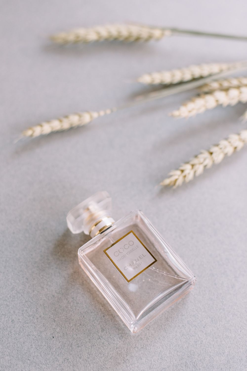 Crystal perfume bottle with a gold lid photographed in minimalist style with dried wheat stalks on a neutral gray background.