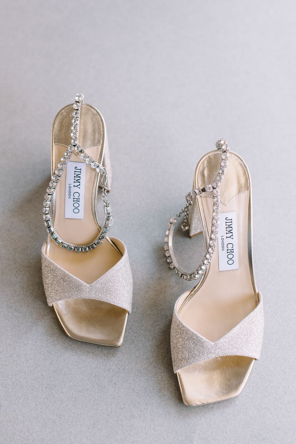 Elegant Jimmy Choo wedding sandals in glittery champagne with crystal-embellished ankle straps on grey background.