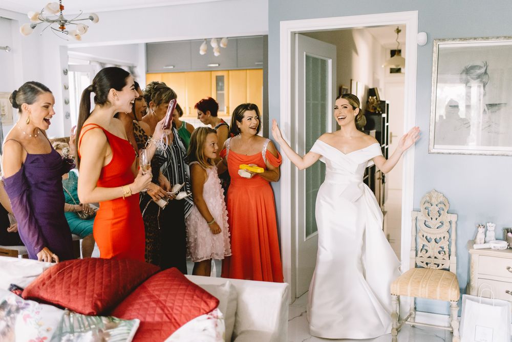 A bridal party gathers in a light blue room with bridesmaids wearing vibrant orange dresses beside someone in a white gown.
