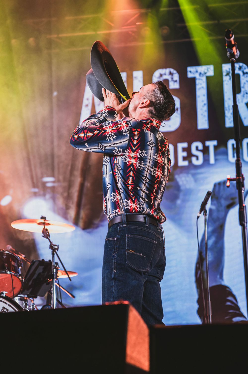 Country music performer in patterned western shirt holding cowboy hat on stage with colorful concert lighting.