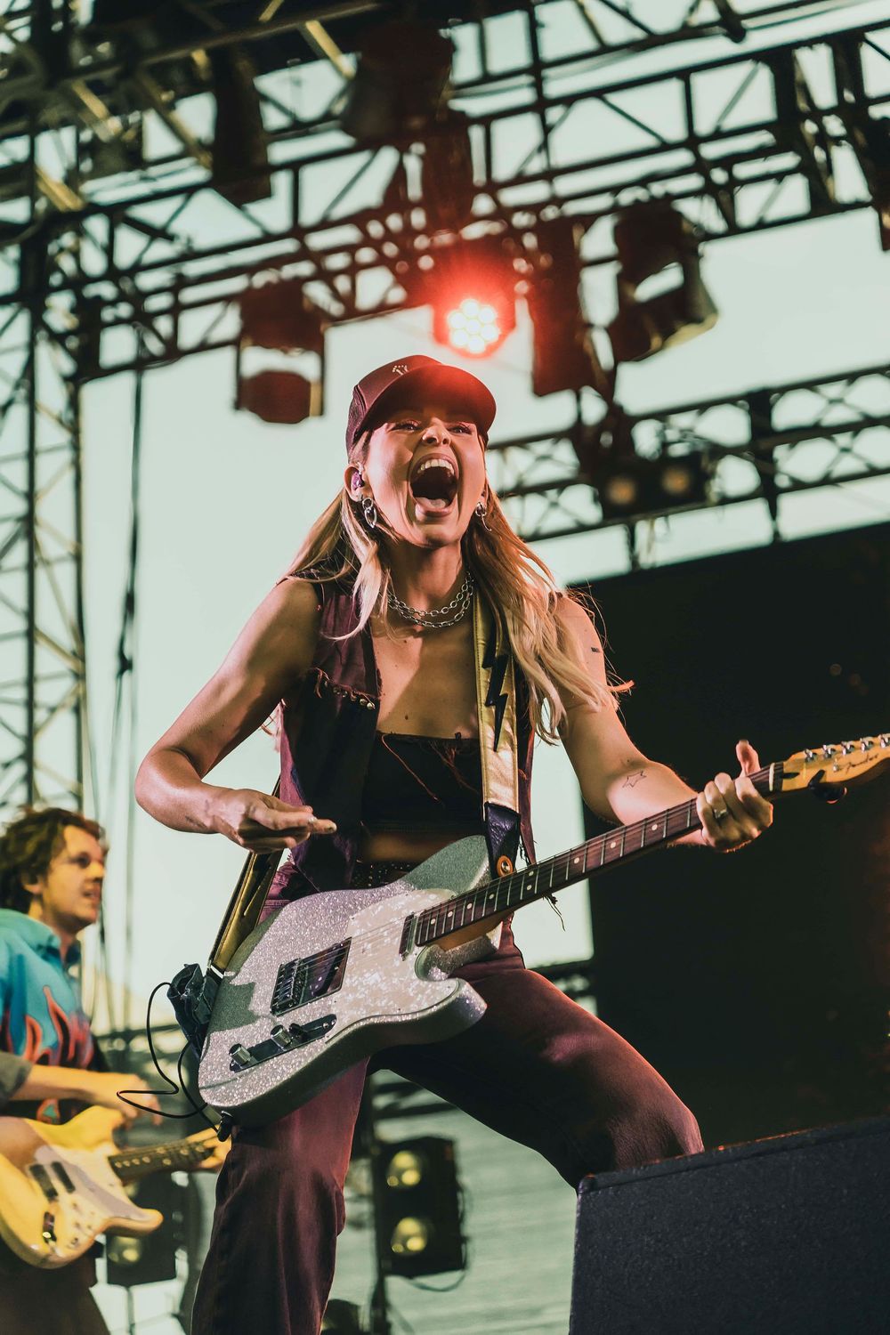 Female guitarist performing energetically on stage at outdoor music festival, playing electric guitar under bright stage lights.