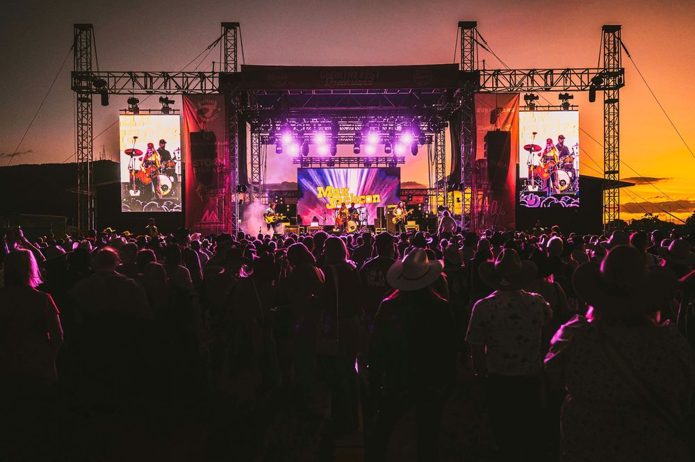 Large crowd enjoys an outdoor music festival at sunset with a brightly lit stage and performers visible in the background.