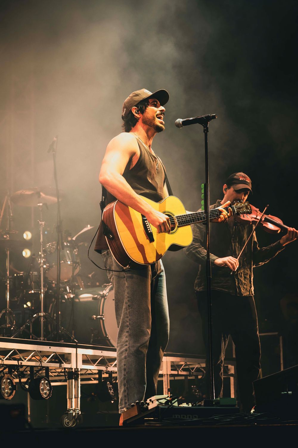 Male guitarist performs on stage with a violin player behind him, both wearing caps under dramatic concert lighting.
