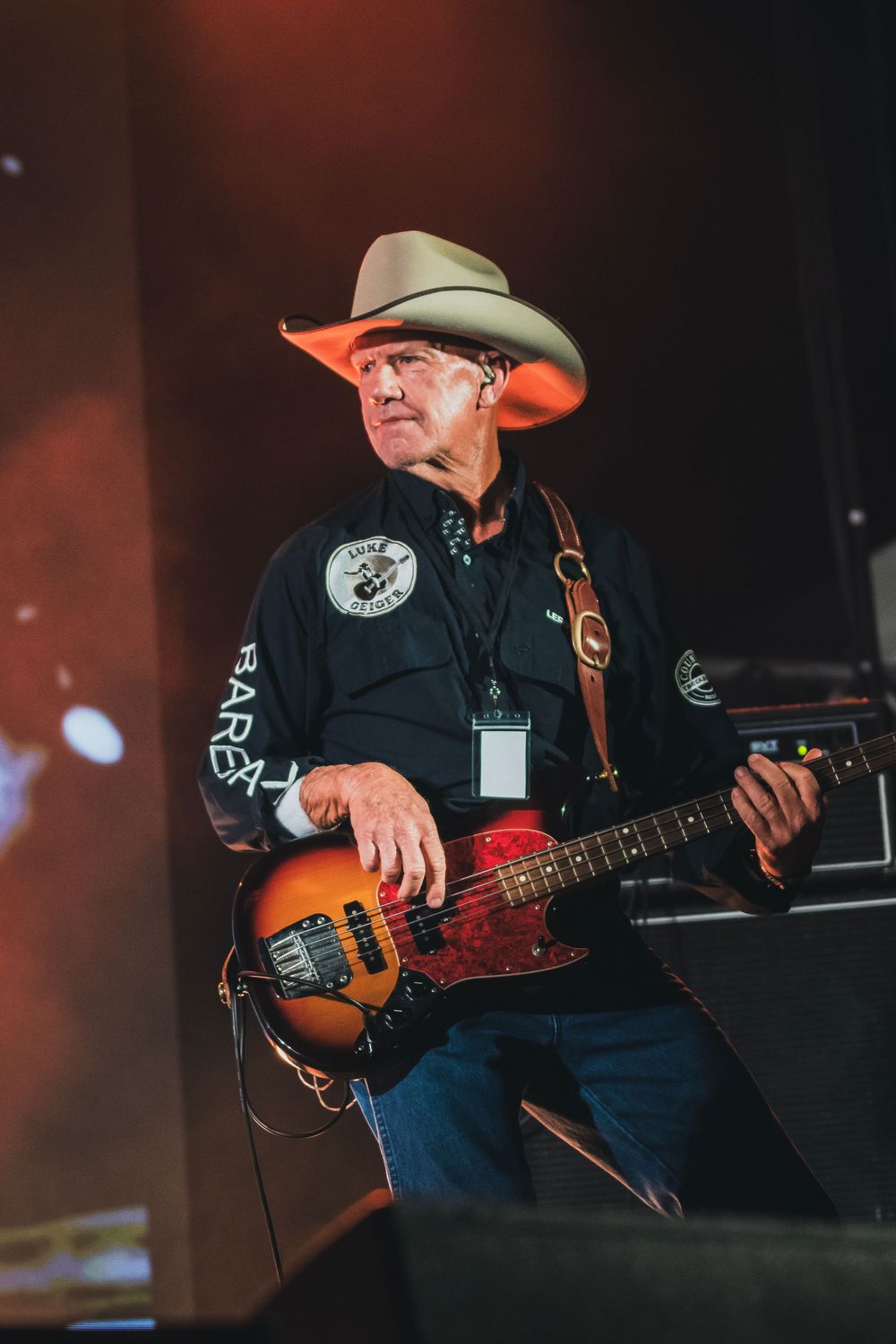 Country musician in a cowboy hat plays a sunburst bass guitar on a warmly lit outdoor stage.