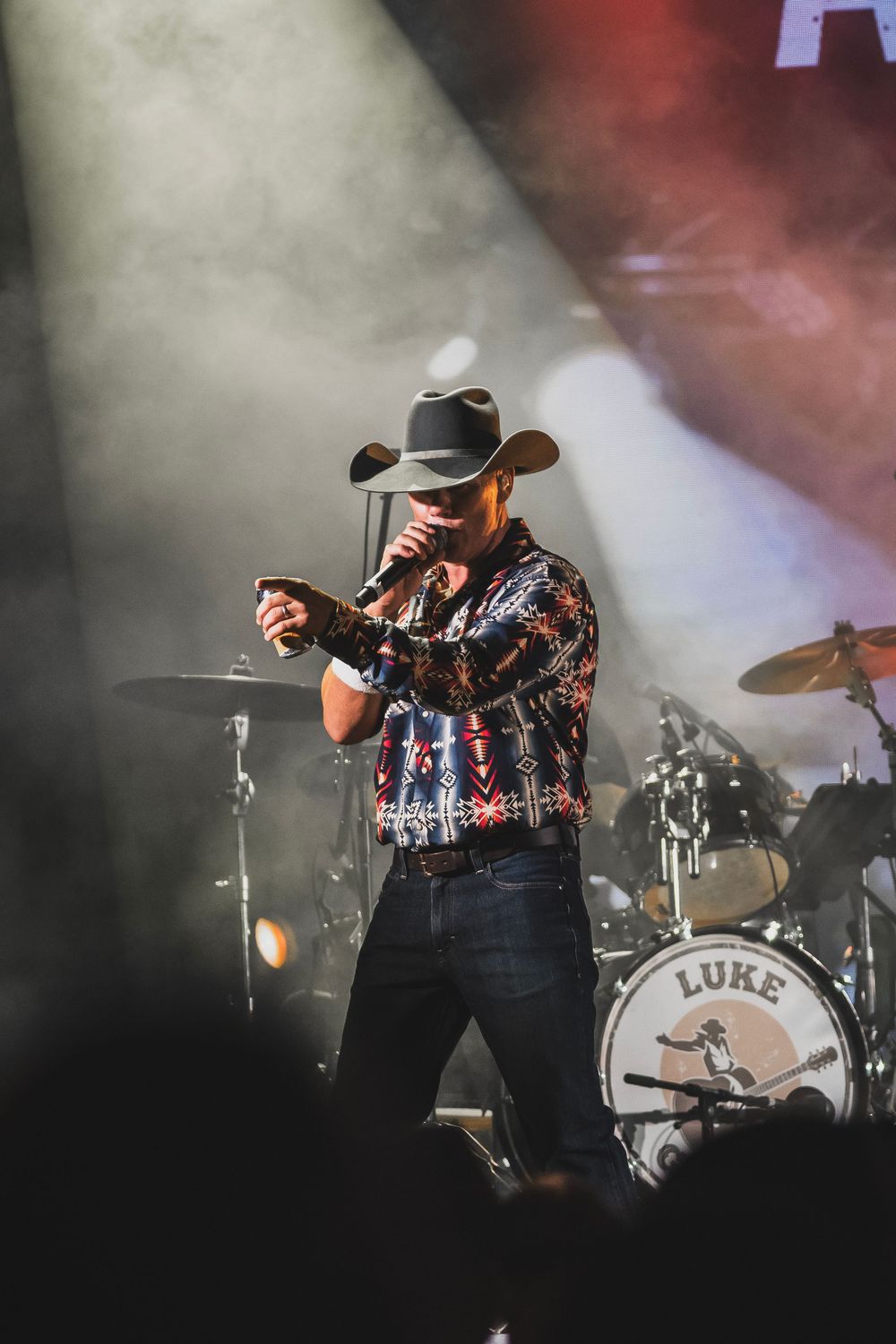 Country singer performing on stage wearing a cowboy hat and patterned shirt, with a drum kit labeled Luke visible behind him.