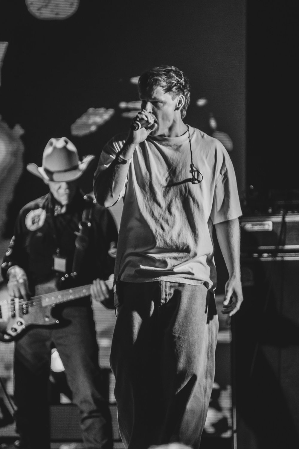 Black and white photo of a male singer performing on stage with a band, holding a microphone in a casual t-shirt.