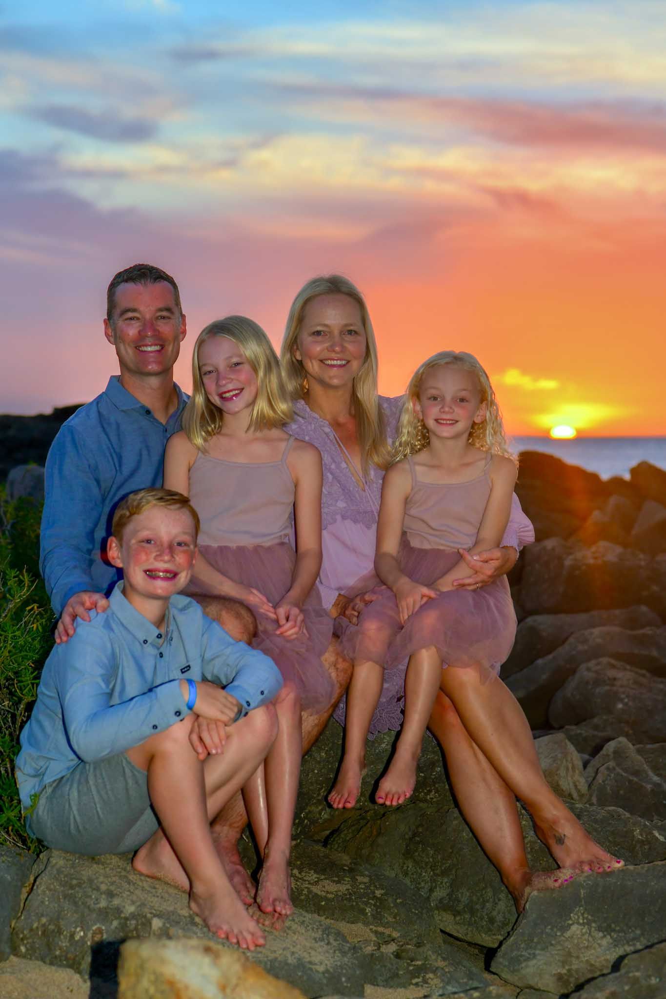 Family shoot session at Ko Olina lagoon poses together on coastal rocks at sunset with a vibrant orange and purple sky in the background.