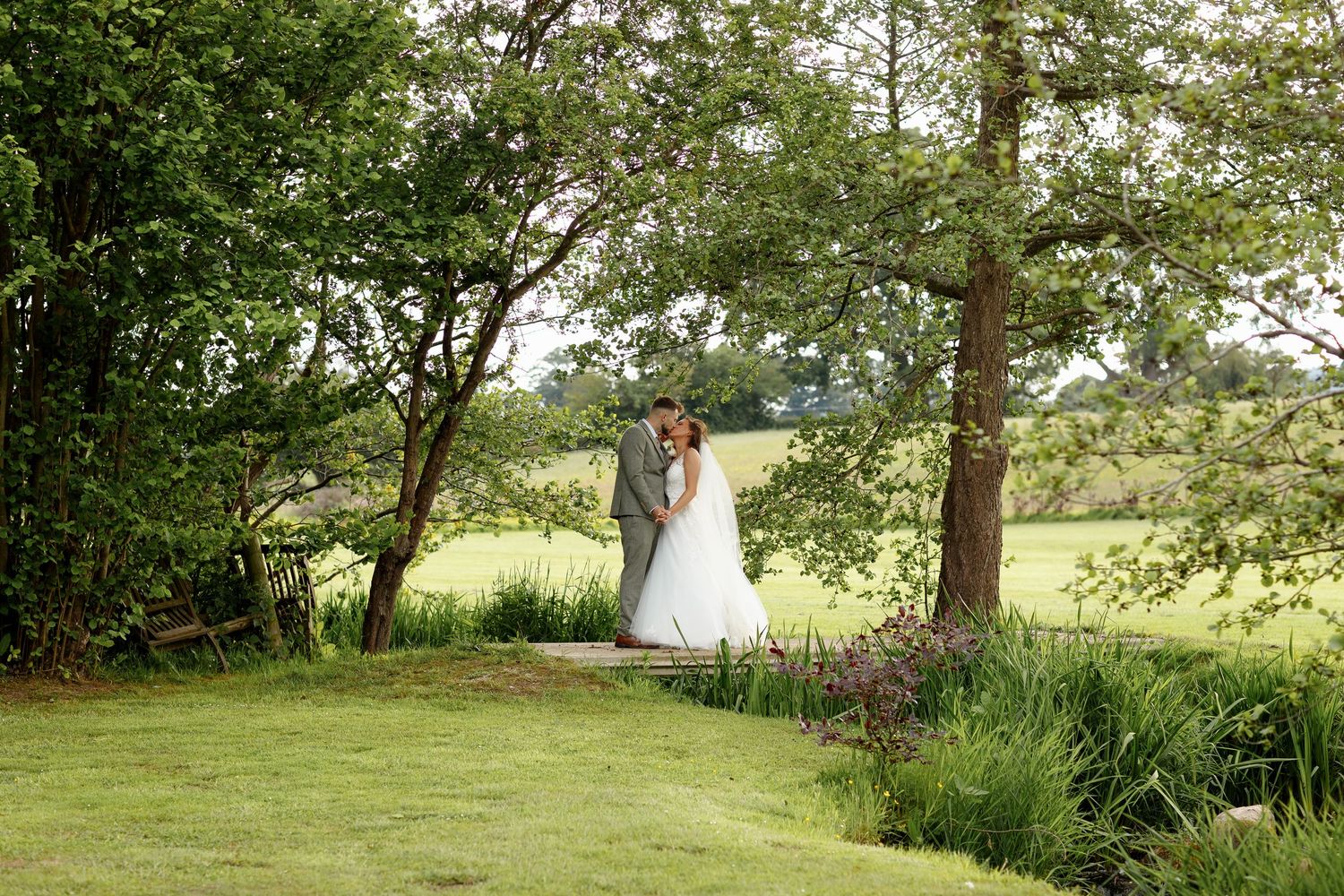 Two figures embrace in an idyllic garden setting surrounded by lush trees and flowering plants on a sunny summer day.