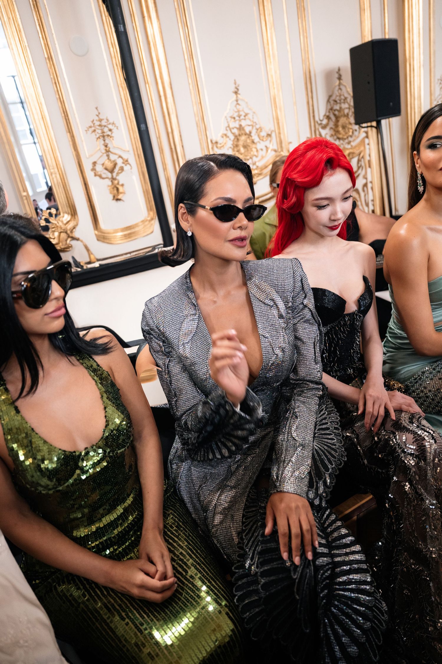 Glamorous guests in sequined dresses and sunglasses seated in an ornate room with gold decor for a luxury event.